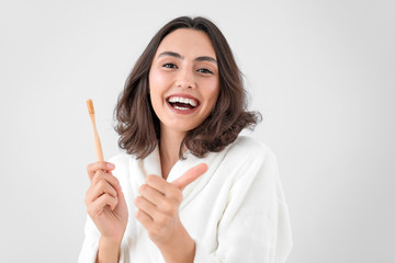 Beautiful young woman with toothbrush showing thumb-up gesture on light background