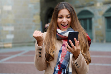 Happy excited young woman laughs watching good news on mobile phone with fist up in city street,...