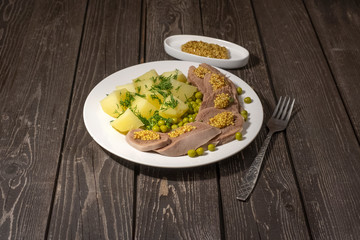 boiled beef tongue with potatoes, green peas, herbs and mustard on dark wooden background with fork
