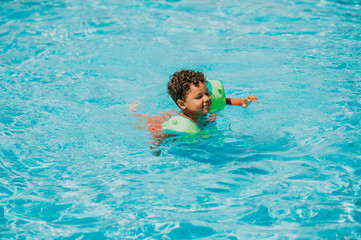 Fototapeta na wymiar Cute african toddler boy swimming in pool with inflatable arm rings