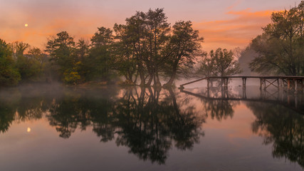 Autumn scenic view of trees and bridge and their reflection during colorful and misty morning sunrise and moonset 