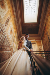 stylish wedding couple of pretty bearded groom and elegant blonde woman in wedding dress posing for photo in chic palace, copy space, noise effect