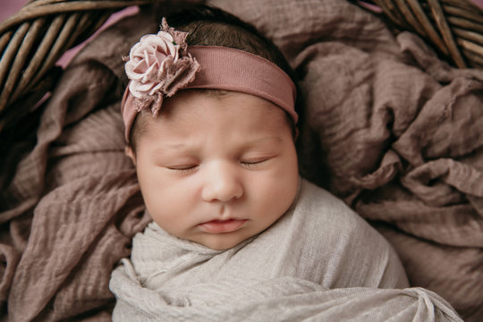 Close up of newborn Infant baby girl swaddled and wrapped in a tan neutral blanket wrap with a dusty pink floral headband on
