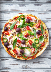 Pizza with cheese, cherry tomatoes, jalapeno pepper, kalamata olives, champignon mushrooms, fresh basil and rocket. Bright wooden background. Top view.	