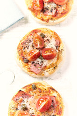 Mini pizzas with ham, cherry tomatoes, mozzarella and capers on bright wooden background. Top view. 