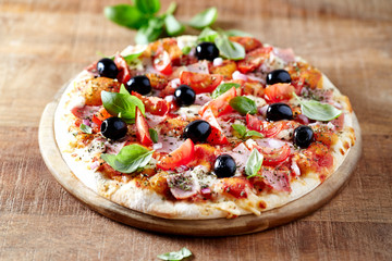 Rustic pizza with ham, cherry tomatoes, black olives and fresh basil on wooden background. 