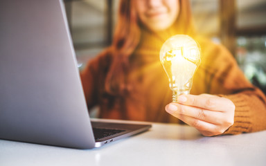Closeup image of a businesswoman holding and showing a glowing light bulb while working on laptop