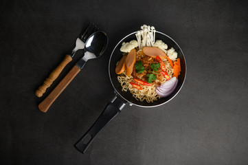 Still life with the noodles in a frying pan, fork, and spoon on the black cement floor.