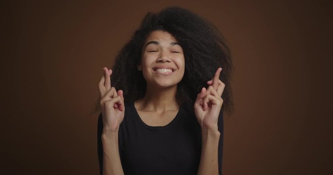 African american girl holding finers crossed, make wish against brown background