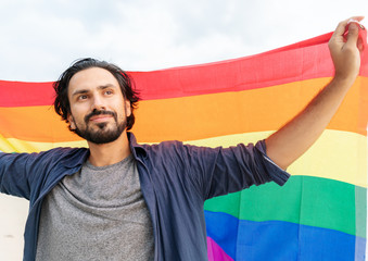 Cheerful guy with a rainbow flag on the beach. Young man holding a rainbow flag against the ocean...