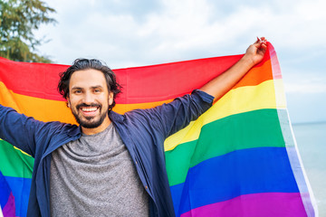 Cheerful guy with a rainbow flag on the beach. Young man holding a rainbow flag against the ocean...