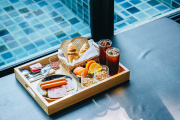 Hand breakfast set with bread, sausage, fruit, ham, yoghurt and coffee served in a wooden tray with a backdrop by the pool
