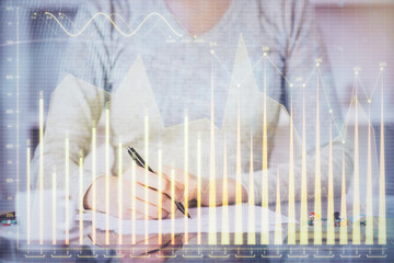 Hand taking notes in notepad. Forex chart holograms in front. Concept of research. Multi exposure