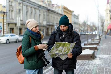 A couple of tourists in the historical center of the city are reading a map while visiting the city and having fun. Girl in winter clothes with camera and cup of coffee and man in winter clothes.