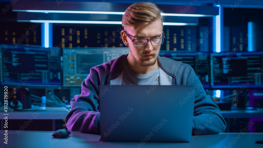 Poster Portrait of Software Developer / Hacker / Gamer Wearing Glasses and Headset Sitting at His Desk and Working / Playing on Laptop. In the Background Dark High Tech Environment with Multiple Displays.