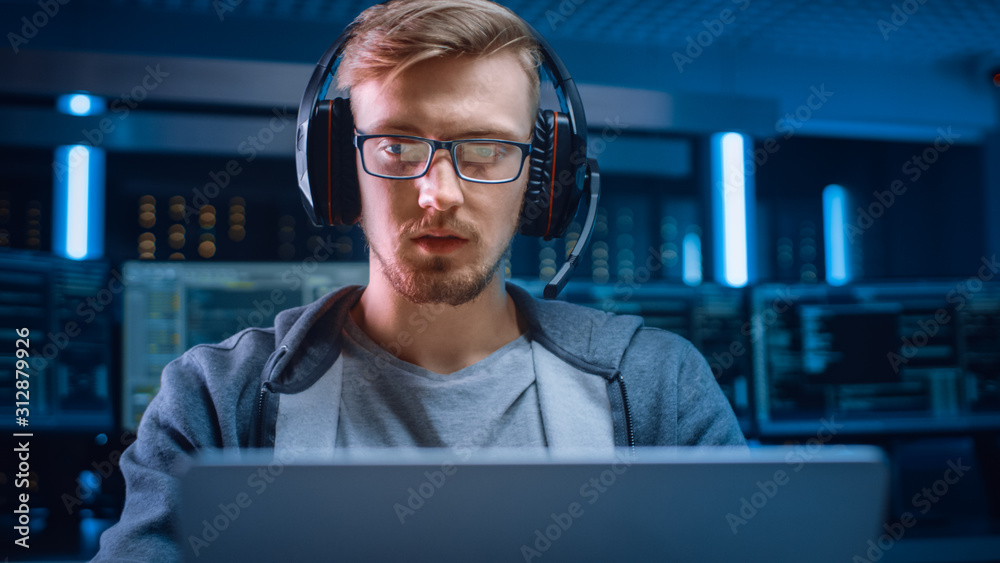 Poster Portrait of Software Developer / Hacker / Gamer Wearing Glasses and Headset Sitting at His Desk and Working / Playing on Laptop. In Background Dark Neon High Tech Environment with Multiple Displays.