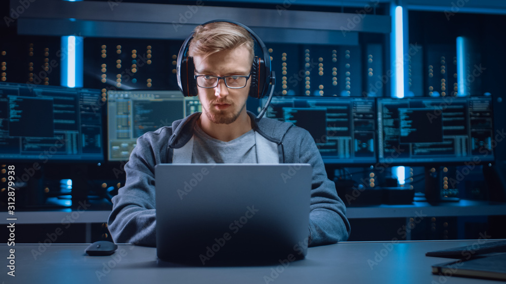 Poster Portrait of Software Developer / Hacker / Gamer Wearing Glasses and Headset Sitting at His Desk and Working / Playing on Laptop. In Background Dark Neon High Tech Environment with Multiple Displays.