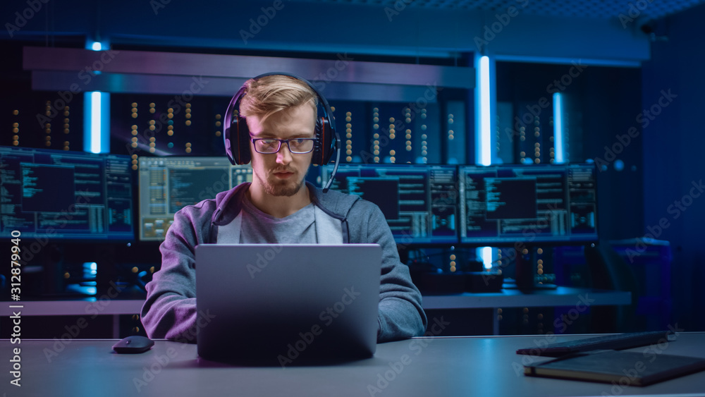 Poster Portrait of Software Developer / Hacker / Gamer Wearing Glasses and Headset Sitting at His Desk and Working / Playing on Laptop. In Background Dark Neon High Tech Environment with Multiple Displays.