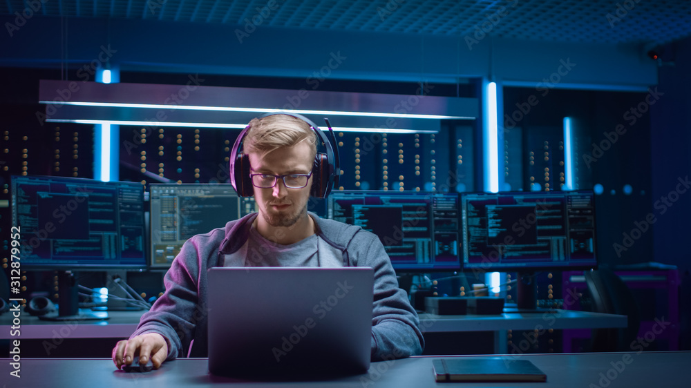 Poster Portrait of Software Developer / Hacker / Gamer Wearing Glasses and Headset Sitting at His Desk and Working / Playing on Laptop. In Background Dark Neon High Tech Environment with Multiple Displays.