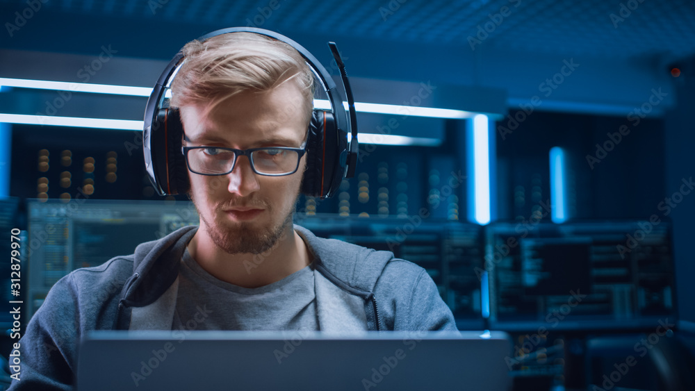 Poster Portrait of Software Developer / Hacker / Gamer Wearing Glasses and Headset Sitting at His Desk and Working / Playing on Laptop. In Background Dark Neon High Tech Environment with Multiple Displays.