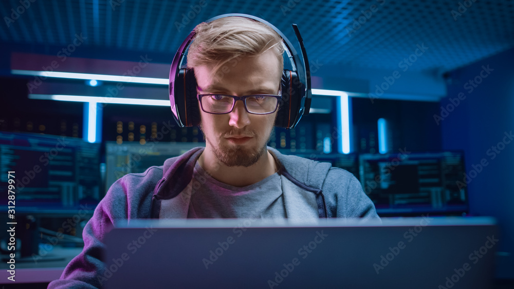 Poster Portrait of Software Developer / Hacker / Gamer Wearing Glasses and Headset Sitting at His Desk and Working / Playing on Laptop. In Background Dark Neon High Tech Environment with Multiple Displays.