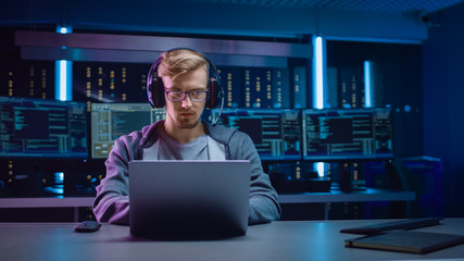 Portrait of Software Developer / Hacker / Gamer Wearing Glasses and Headset Sitting at His Desk and Working / Playing on Laptop. In Background Dark Neon High Tech Environment with Multiple Displays.