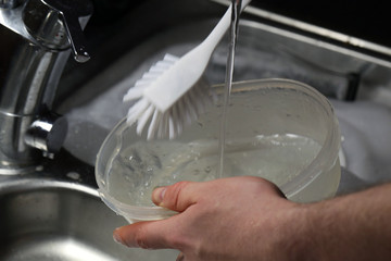Doing dishes after cooking at home. Caucasian male doing the dishes. Closeup image. In this photo there is a transparent box being washed with a lot of soup bubbles and running water. Color image.