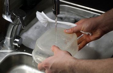 Doing dishes after cooking at home. Caucasian male doing the dishes. Closeup image. In this photo there is a transparent box being washed with a lot of soup bubbles and running water. Color image.