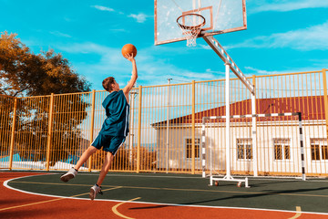 Sports and basketball. A young teenager in a blue tracksuit jumps and throws a ball into the basket. Blue sky and school in the background