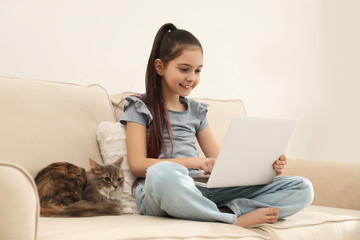 Cute little girl with laptop and cat on sofa at home. First pet
