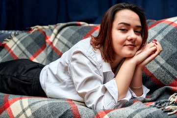 Portrait of a lovely brunette caucasian girl in a white shirt at home on the sofa. Lies and relaxes on a plaid in front of the camera in various poses.