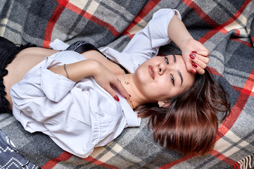 Naklejka premium Portrait of a lovely brunette caucasian girl in a white shirt at home on the sofa. Lies and relaxes on a plaid in front of the camera in various poses.