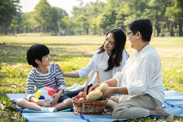 Asian family having picnic in fresh green park together. Mother talking to kid with smile while grandmother preparing food in basket. Cute child looking at mom with happiness. Relax family activity.