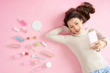 Obraz na płótnie Canvas Young Asian woman taking selfie with her phone and lying on pink background with her cosmetics. Top view