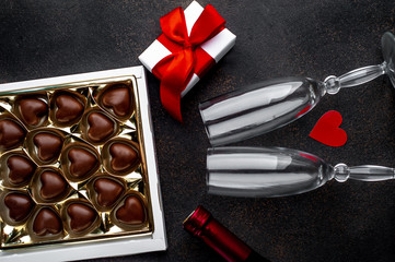 Heart shaped candies in box, gift with red  ribbon, a bottle of wine or champagne and two glasses for Valentine's Day on a stone background with copy space for your text. Valentine's Day celebration 
