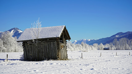 a wood hut on a agriculture field in winter with fresh snow and a blue clear sky with view to the mountains