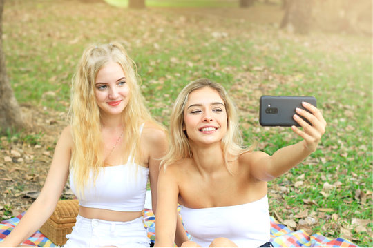 Happy friends on picnic in park, two young beautiful teen girls taking selfie with smartphone and having fun while spending time together at summer garden, woman outdoor friendship concept.
