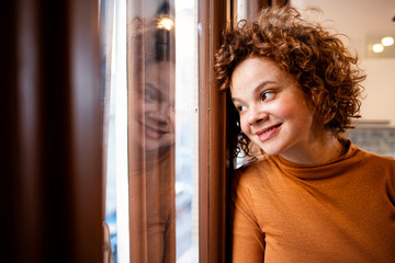 Young woman looking out window, view through glass, close-up. Beautiful woman at the window. Beautiful young brunette woman looking relaxed at home.
