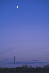Picture of a field at twilight. Blue evening sky with the moon. High voltage powerline above the trees.