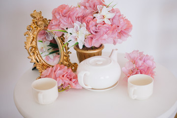 Tea table, mugs and a teapot white teapot with a pink bouquet of flowers in the rococo style. Golden vintage mirror. White pink eltion interior