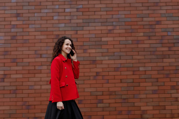Outdoors portrait of a business woman in red, hair in wind, talking on the smartphone while walking outside, near brick wall. woman happy using mobile phone app for conversation. Space for text.