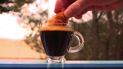 A man dunks a fresh croissant in hot coffee in a transparent cup on the railing of the balcony against the background of nature in the early morning. - Powered by Adobe