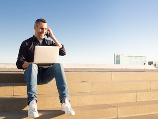 Handsome middle aged business man talking on the phone while using laptop sitting on stairs at daytime - Powered by Adobe