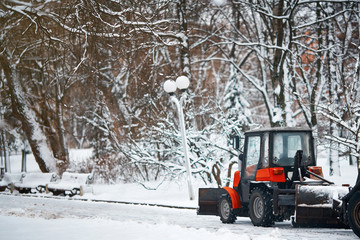 Red small tractor clearing snow in central park, with snow plow and rotating brush. Municipal...