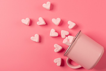 a pink mug lies on its side against a pink background, light-pink marshmallows sprinkled from it in the form of hearts