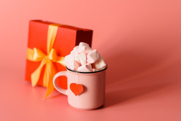 pink mug on a pink background filled with marshmallows in the form of hearts. next to it is a box with a gift for Valentine's Day. valentine's day holiday concept