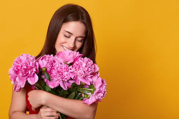 Fototapeta premium Indoor studio shot of tender charming young female having pleasant facial expression, smelling pink peonies, getting pleasure of presents, being in high spirits. Copyspace for advertisement.