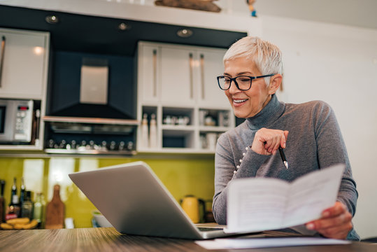 Happy Woman Looking At Laptop, Holding Documents And Sitting In The Kitchen, Portrait.