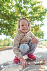 little boy 3-5 years old, squats, draws drawing with colored crayons asphalt. Resting weekend, walk in autumn in park. Happy laughs and smiles. Warm casual wear, beige sweater with a hood.