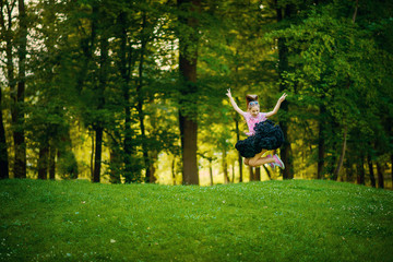 A beautiful girl in a fluffy black skirt tutu joyfully jumps on a green lawn among the trees. The concept of happy childhood.